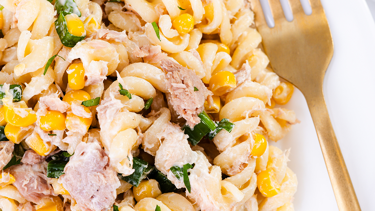 A close-up of a pasta salad with spiral noodles, chunks of tuna, corn, green herbs, and creamy dressing, served on a white plate with a gold fork—perfect for easy Memorial Day Recipes.