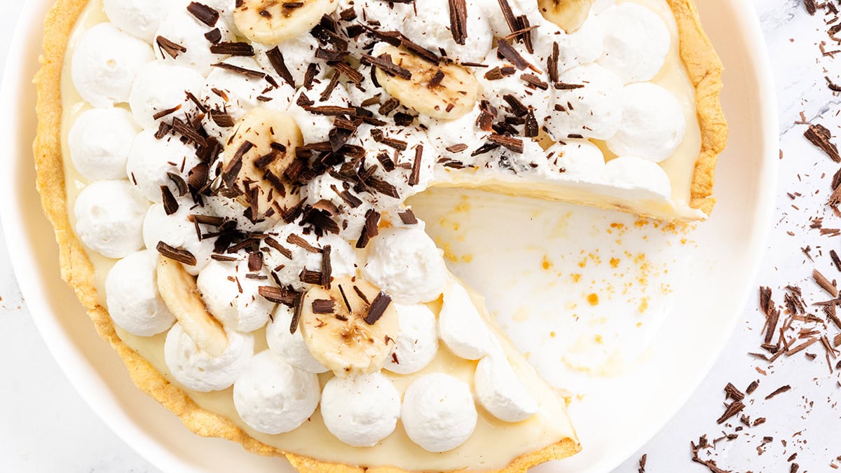 An easy dessert, this banana cream pie is topped with whipped cream, banana slices, and chocolate shavings, with one slice removed to reveal its creamy filling and golden crust.
