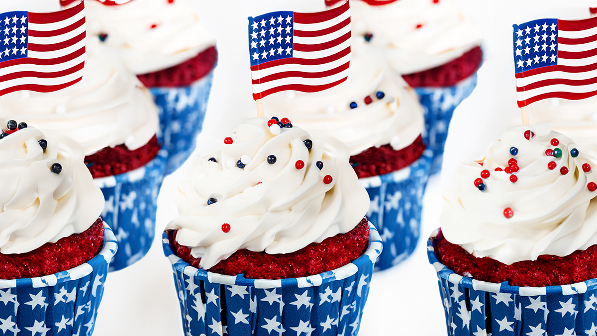 Red velvet cupcakes with white frosting, decorated with red, white, and blue sprinkles and miniature American flags—perfect Memorial Day recipes. The cupcake wrappers have a blue background with white stars.