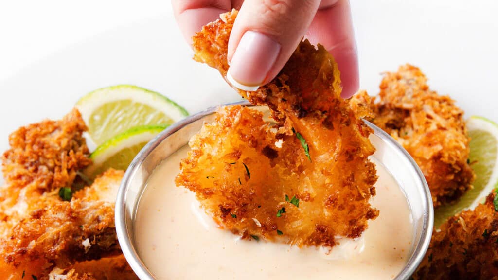 A hand dips a crispy, golden-fried shrimp into a creamy dipping sauce, with lime slices and more fried shrimp on a plate in the background—perfect for your next round of picnic recipes.