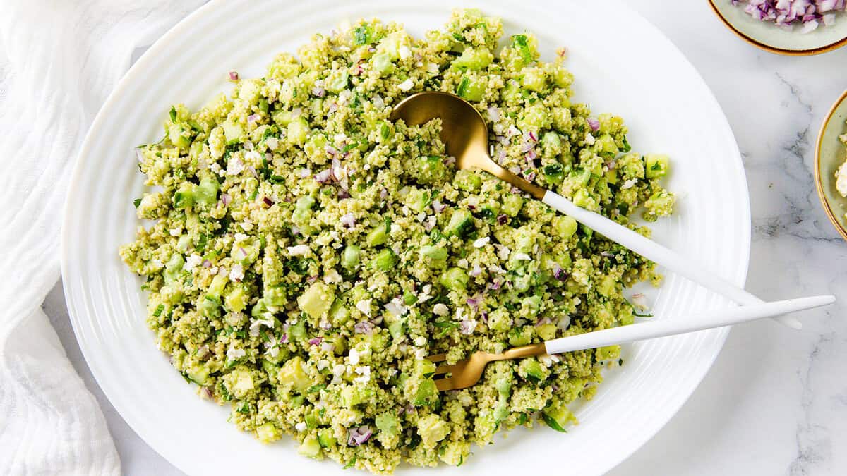 Piring putih besar yang diisi dengan salad quinoa hijau, menampilkan sayuran dan rempah -rempah cincang, menampilkan salah satu resep salad segar. Dua peralatan melayani bertumpu di atas. Di latar belakang adalah kain putih dan piring kecil.