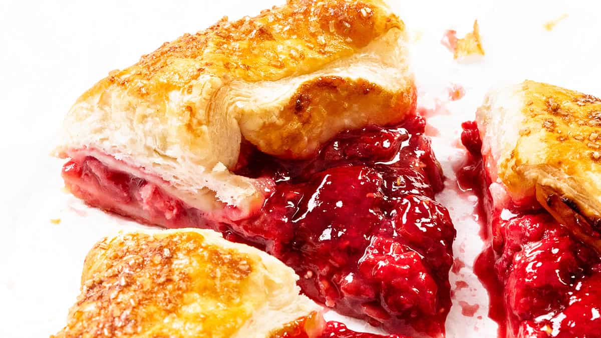 Close-up of a flaky puff pastry with golden crust and sugar crystals, filled with bright red, juicy berry filling that is oozing out. Perfect for those who love creative Puff Pastry Recipes, the pastry is cut into pieces on a white surface.