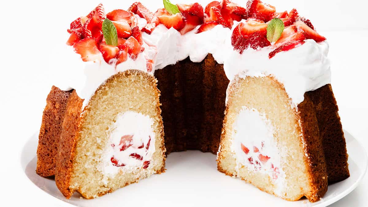 A sliced bundt cake filled with whipped cream and strawberries, topped with more whipped cream, strawberry pieces, and mint leaves—a classic among strawberry desserts—displayed on a white plate against a white background.