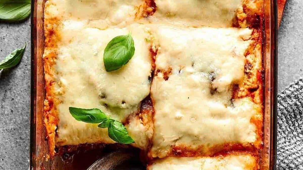 A close-up of a baked lasagna in a glass dish, topped with melted cheese and fresh basil leaves. Perfect for fans of Eggplant Recipes, this dish is ready to serve with a spoon in one corner and a textured cloth beside it.