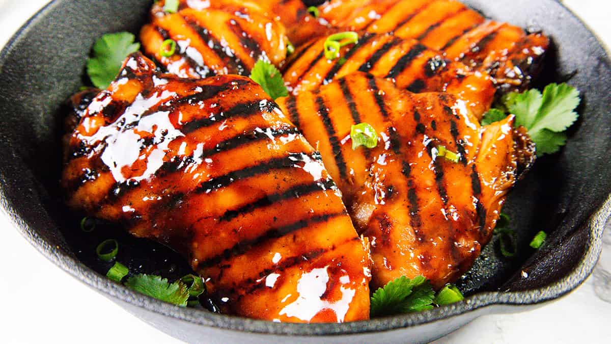 Paha ayam bbq asian lengket bakar dengan bekas panggangan gelap dalam wajan hitam, dihiasi dengan bawang hijau dan herbal segar