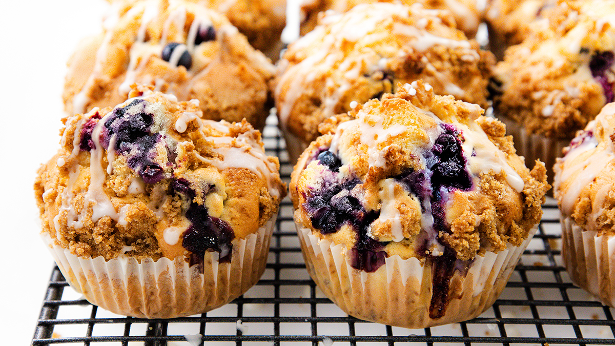 Freshly baked blueberry muffins with crumb topping and glaze, arranged on a cooling rack—a scrumptious breakfast to kickstart your day. The muffins are golden brown with juicy blueberries and drizzled icing.