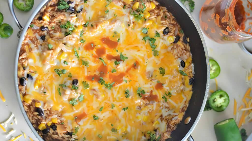 A close-up shot of a pan filled with a cheesy Mexican rice dish perfect for easy dinners. The rice is topped with melted cheese, black beans, corn, chopped cilantro, and drizzled with hot sauce.