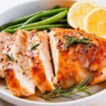 A plate of sliced, Slow Cooked Crockpot Chicken Breast garnished with rosemary sprigs. The chicken is accompanied by fresh green beans and two lemon slices, all arranged on a white plate. The chicken has a golden-brown, seasoned exterior.