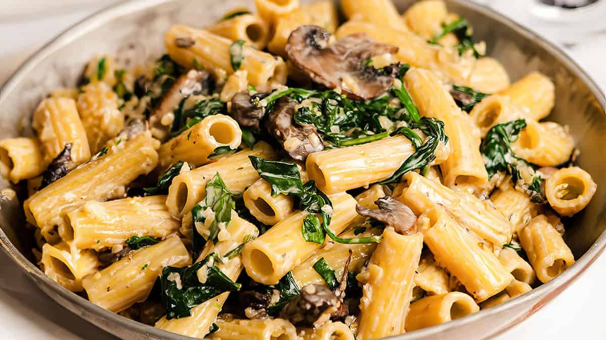 A close-up of creamy rigatoni pasta mixed with sautéed spinach and sliced mushrooms, served in a large silver pan—perfect for anyone searching for comforting pasta recipes.