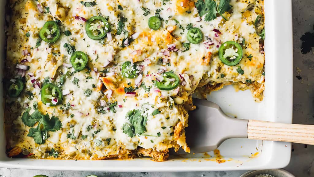 A rectangular white dish of mouth-watering Salsa Verde Chicken Casserole is shown, topped with melted cheese, sliced jalapeños, chopped red onions, and cilantro.