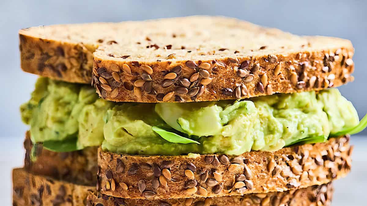 Tampilan jarak dekat dari sandwich yang dibuat dengan dua potong roti multigrain. Isiannya berupa alpukat tumbuk dan daun bayam segar, dengan biji yang terlihat jelas di kulit roti. Sempurna untuk sarapan atau makan siang, latar belakangnya sedikit kabur, memusatkan perhatian pada sandwich yang lezat.