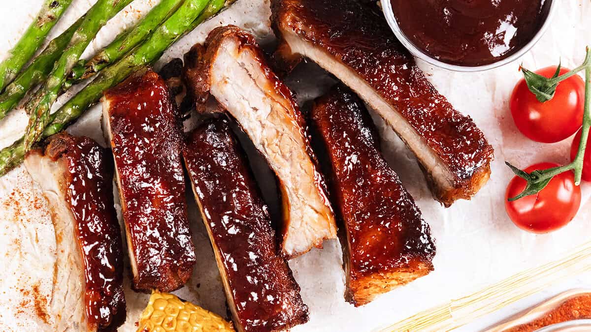 Barbecue ribs with a shiny glaze, inspired by top bbq recipes, served with grilled asparagus, cherry tomatoes, corn on the cob, and a cup of barbecue sauce on a white surface.