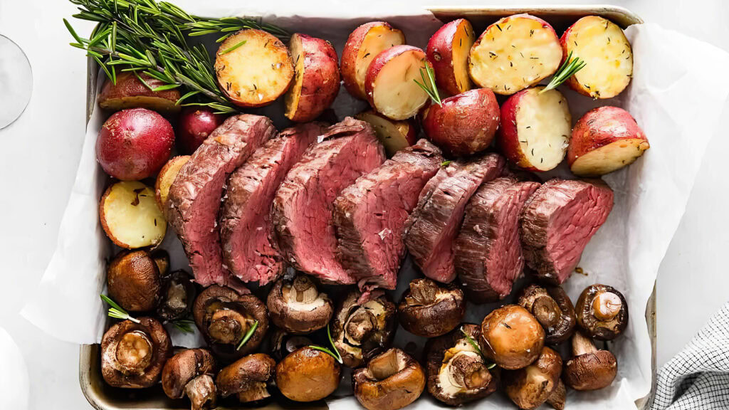 A tray with evenly sliced medium-rare roast beef in the center, surrounded by halves of roasted red potatoes garnished with rosemary, and a variety of roasted mushrooms. This delicious spread is an excellent addition to your sheet pan recipes collection. The tray is lined with parchment paper.