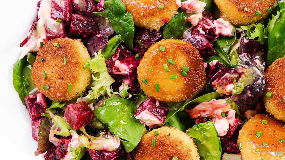 Tampilan dekat salad dengan sayuran hijau, bit potong dadu, keju putih krem, dan putaran keju kambing berlapis-pantat keemasan, dihiasi dengan daun bawang cincang-tambahan yang lezat untuk resep salad favorit Anda.