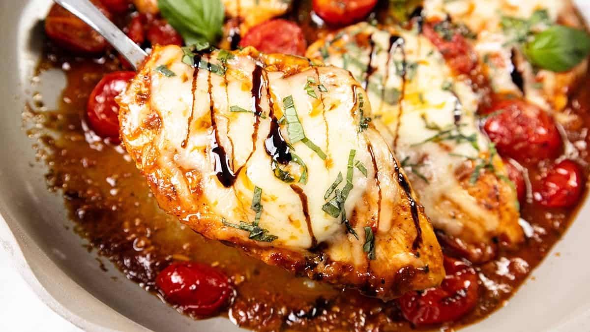 A serving of chicken breast topped with melted cheese, chopped basil, and a drizzle of balsamic glaze, surrounded by roasted cherry tomatoes and sauce in a pan—a standout dish for lovers of Cheesy Recipes.