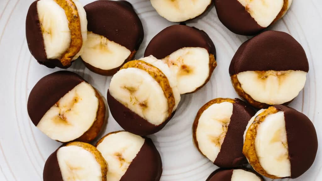 A plate of bite-sized snacks, each consisting of a banana slice sandwiched with a layer of nut butter and partially dipped in dark chocolate. Perfect for those exploring new snack recipes, these treats are arranged in a random pattern on a white surface.