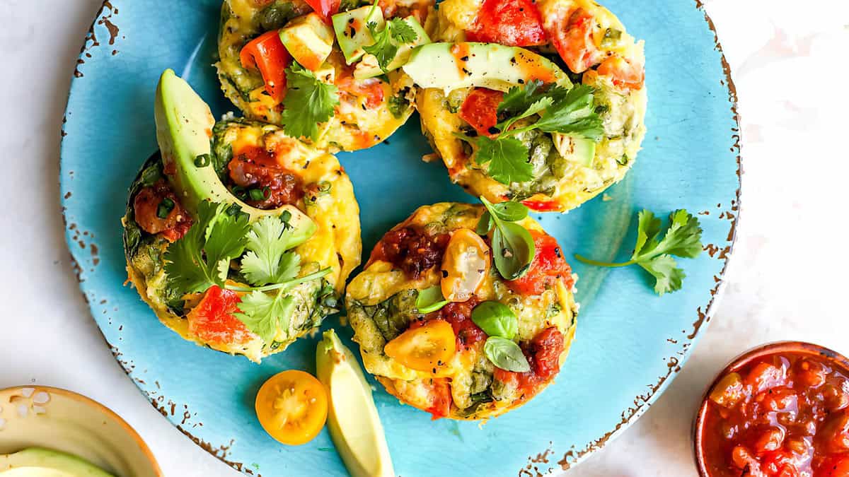 Piring sarapan yang semarak dengan empat frittata sayuran mini yang dihias dengan irisan alpukat, rempah segar, dan salsa tomat. Frittatasnya berwarna-warni, menampilkan paprika, bayam, dan keju. Di sekeliling piring terdapat piring kecil berisi salsa dan tomat ceri—resep telur mudah yang cocok untuk makan siang atau makan malam juga.
