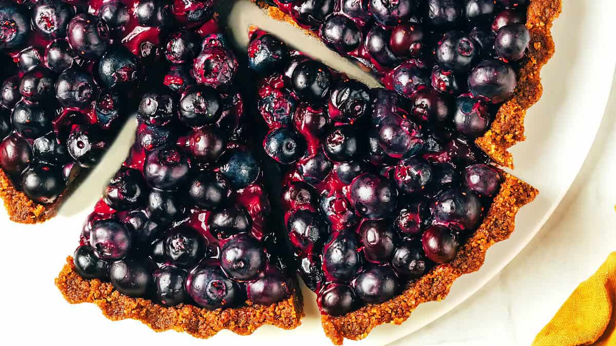 A close-up of a blueberry tart with a few slices cut. The tart has a golden brown crust and is topped with glistening, juicy blueberries. This dessert features vibrant blue and purple hues that contrast beautifully with the golden crust, giving it an appetizing appearance perfect for Sweet Treats.