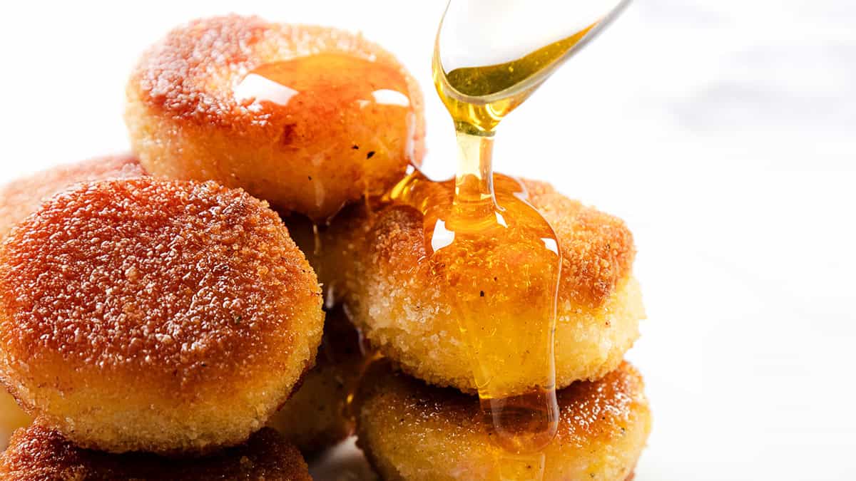 A close-up of a stack of golden brown, Crispy Breaded Fried Goat Cheese being drizzled with honey or syrup. The syrup glistens as it flows over the warm, textured surface—a perfect highlight for fans of golden recipes.