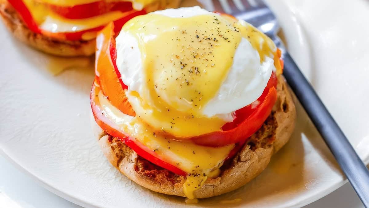 Tampilan jarak dekat dari hidangan yang menampilkan muffin Inggris panggang dengan irisan tomat, telur rebus, dan saus hollandaise di atasnya. Sausnya dihias dengan lada hitam bubuk. Sempurna untuk sarapan atau makan siang ringan dan makan malam, kreasi menyenangkan ini dipadukan dengan garpu di piring putih.