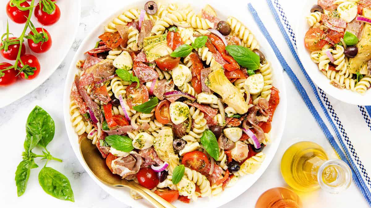 Semangkuk salad pasta Italia dengan rotini, tomat ceri, bola mozzarella, zaitun, artichoke, bawang merah, kemangi, dan daging yang diiris, dihiasi dengan bumbu. Sempurna untuk mereka yang mencari resep salad pasta yang lezat. Tomat dan daun kemangi ada di samping.