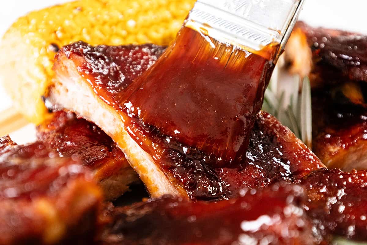 A close-up of barbecue ribs being brushed with a thick layer of glossy sauce, with a corn on the cob in the background—a mouthwatering scene for anyone who loves bbq recipes.