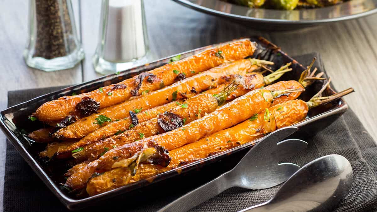 A rectangular black dish filled with roasted whole carrots garnished with herbs, presented as one of the delicious side dishes, sits on a dark napkin beside a spoon, fork, and a salt and pepper shaker.
