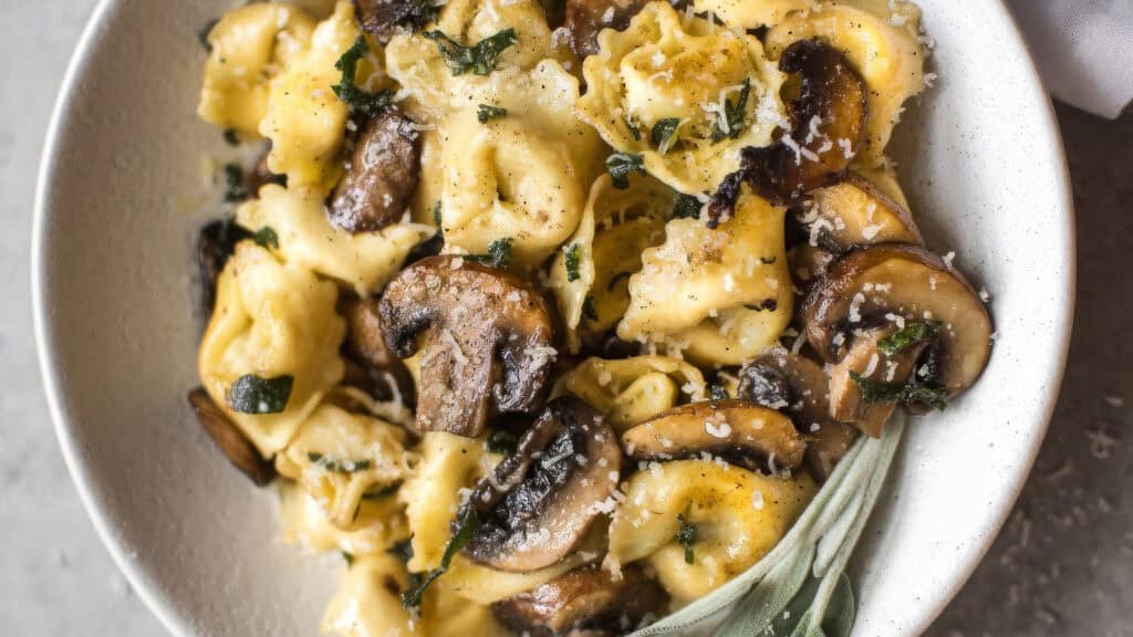A white bowl filled with creamy tortellini pasta mixed with sautéed mushrooms and garnished with grated Parmesan cheese and chopped herbs.