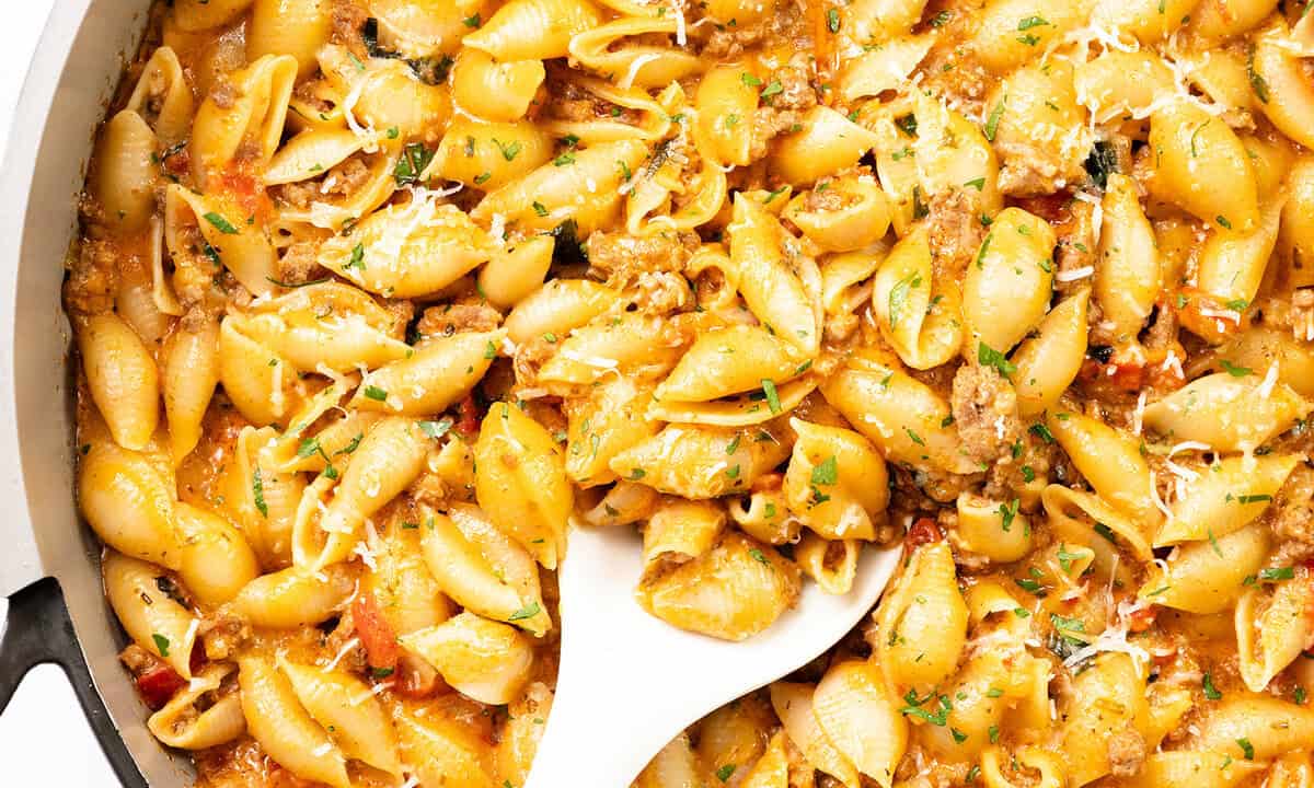 A close-up of creamy pasta shells mixed with ground beef, herbs, and grated cheese, being stirred with a white spoon. This rich and flavorful dish is one of the best ground beef recipes, garnished with chopped parsley.