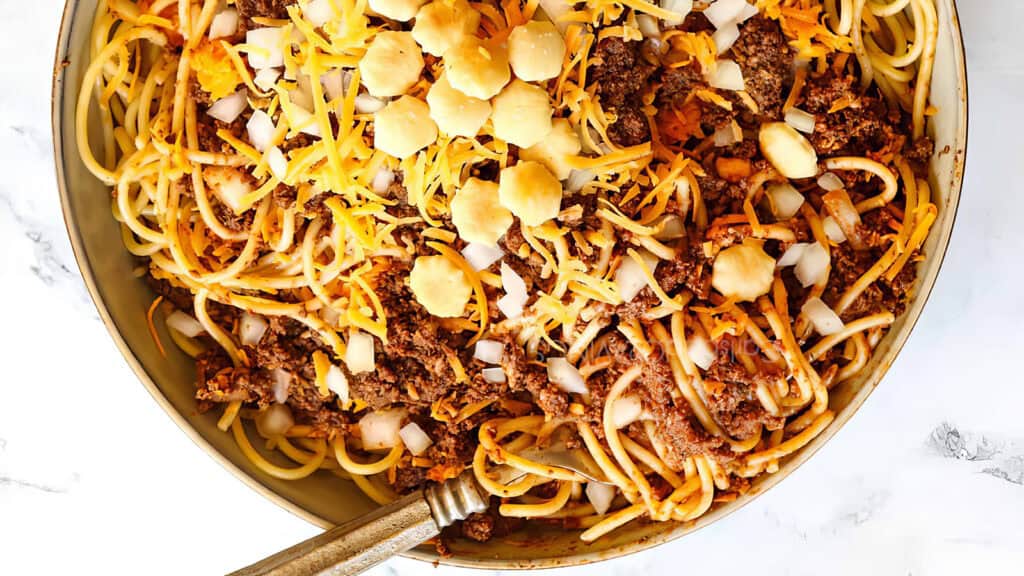 A skillet filled with spaghetti topped with ground beef, shredded cheddar cheese, diced onions, and round oyster crackers rest on a white marble surface.