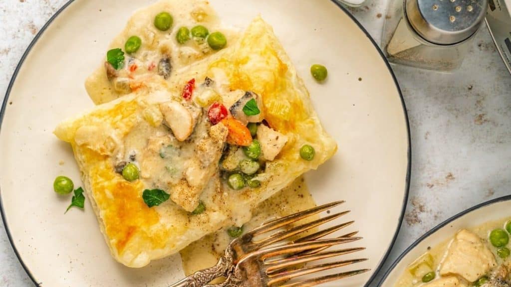 A creamy chicken and vegetable mixture with peas and carrots, inspired by slow cooker recipes, is served on a flaky golden pastry square, garnished with herbs on a white plate with a fork and spoon.