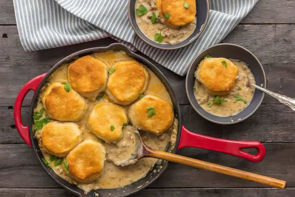 A red skillet filled with creamy sausage gravy topped with golden biscuits, a classic from Southern Recipes, with a portion served in a gray bowl. A striped cloth and silver spoon are nearby on a rustic wooden table.
