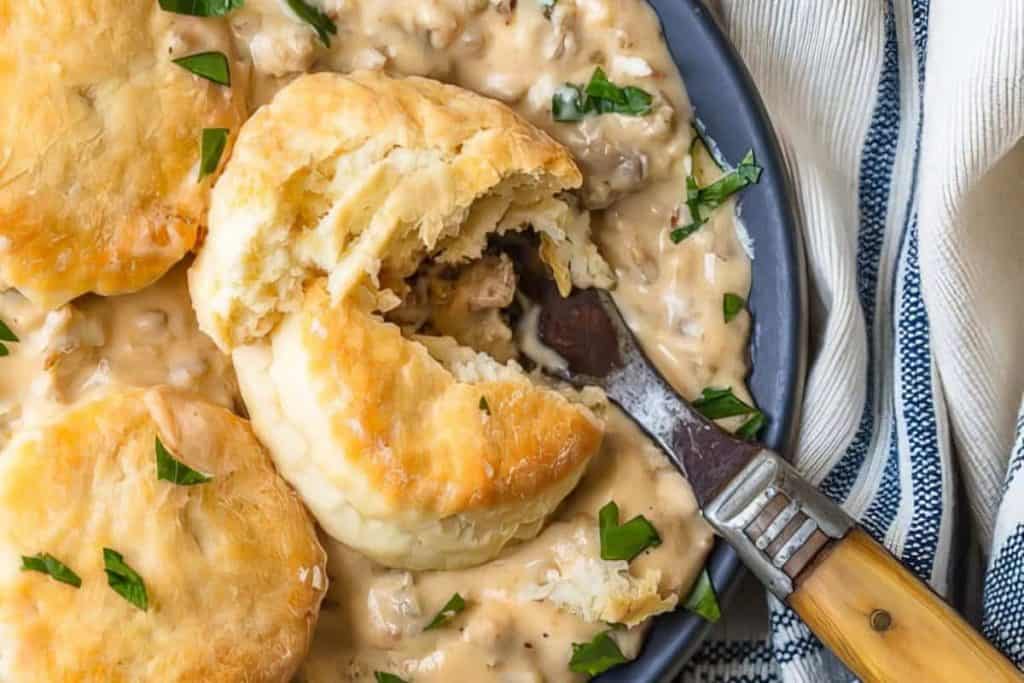 A flaky biscuit sits on a plate covered in creamy sausage gravy, garnished with chopped parsley—a classic from Southern recipes. A fork rests on the plate, breaking open the biscuit, with a striped cloth napkin beside it.