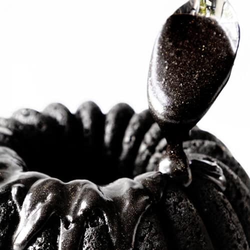 A black velvet bundt cake is being drizzled with shiny black icing glaze from a spoon. The cake's texture is rich and moist, and the smooth glaze adds a glossy finish. The background is a bright white, highlighting the dark hues of the dessert.