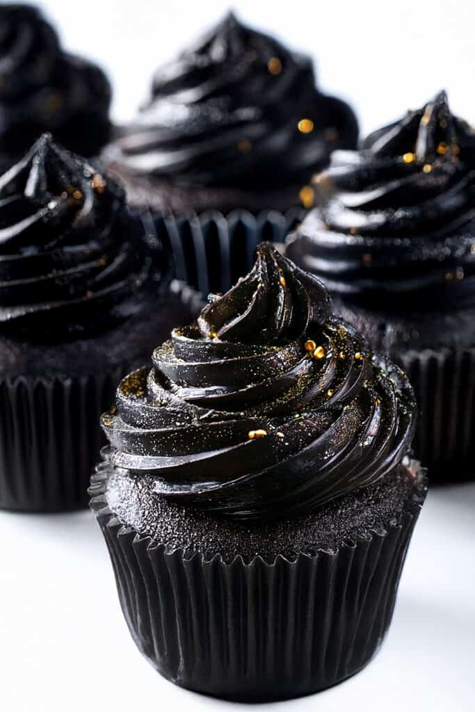 Close-up of Black Velvet cupcakes with glossy dark frosting, accented by small gold sprinkles. The cupcakes are arranged on a white surface, creating a striking contrast.