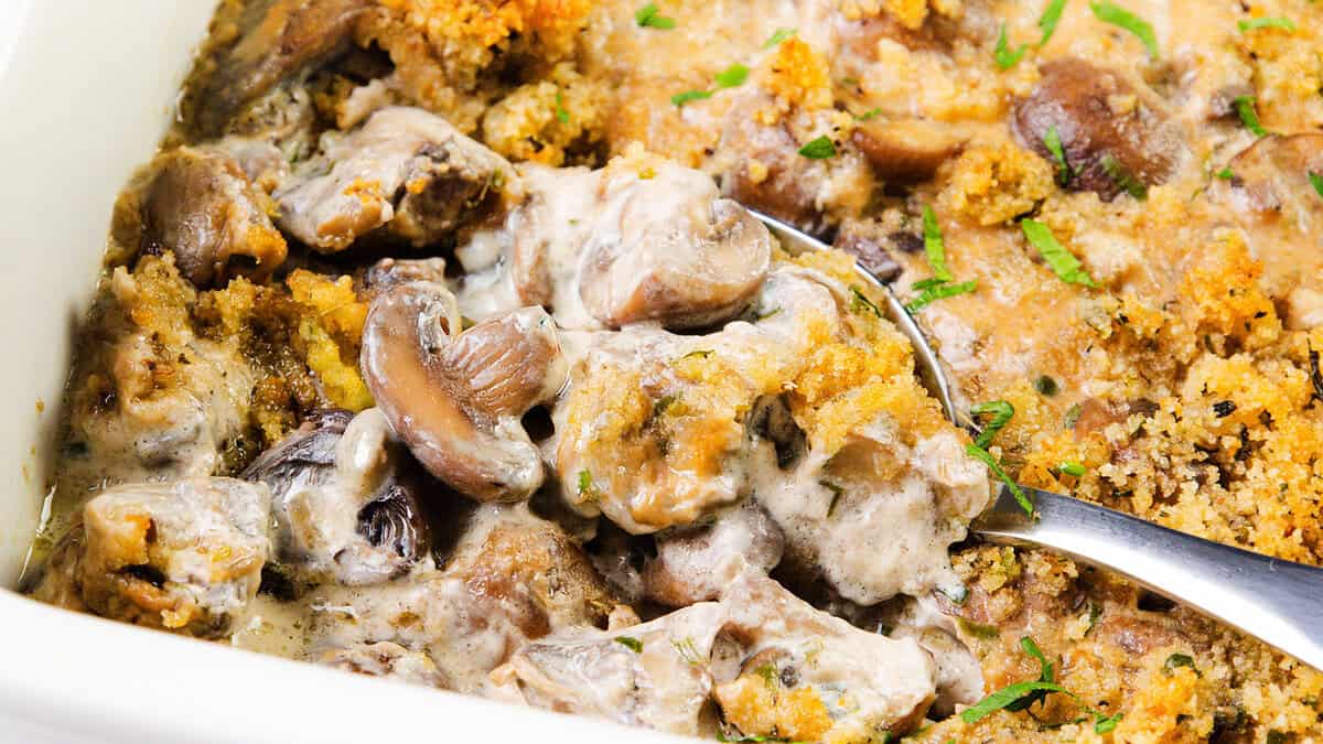 A close-up of a creamy mushroom casserole topped with golden-brown breadcrumbs, served in a white dish with a metal spoon scooping a portion—perfect for Thanksgiving recipes.