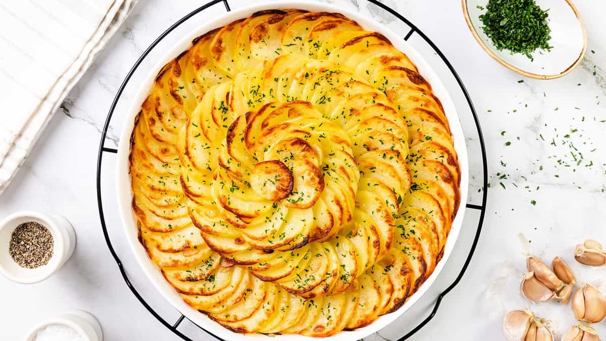 A round white dish filled with beautifully arranged, golden-brown, thinly sliced potatoes in a spiral pattern—an elegant addition to your collection of potato recipes—garnished with chopped parsley. Nearby are seasonings, garlic, and a bowl of herbs on marble.
