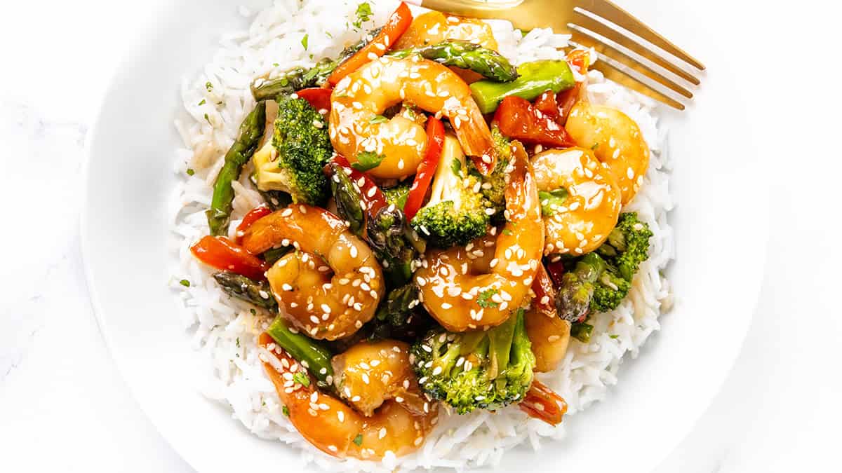 A white plate filled with white rice topped with shrimp, broccoli, asparagus, red bell peppers, and sesame seeds, showcasing a vibrant touch for asparagus recipes, with a gold fork on the side.