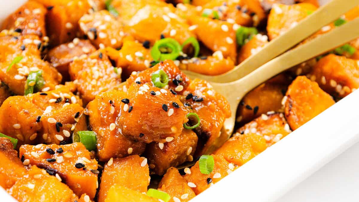 Close-up of roasted sweet potato cubes garnished with sesame seeds and sliced green onions, served in a white dish with a gold serving fork—a delicious addition to your collection of Sweet Potato Recipes.
