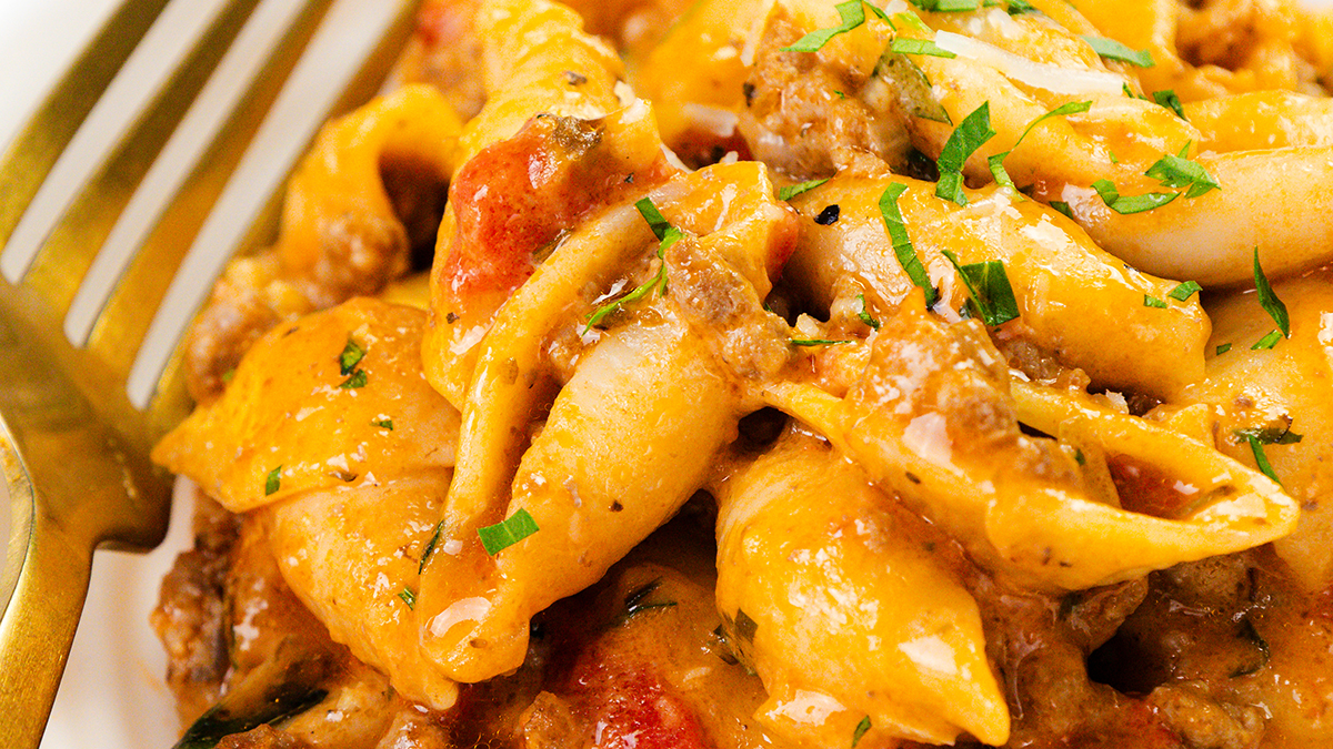 Close-up of creamy pasta shells with ground meat, tomatoes, and melted cheese, garnished with chopped herbs. A gold fork rests next to the pasta on the plate—perfect for easy dinners.