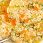 A blue pot filled with lemon chicken orzo soup, brimming with sliced carrots, celery, and fragrant fresh herbs. Lemon wedges add a citrusy twist, while a ladle rests inside this deliciously inviting soup, ready to serve.