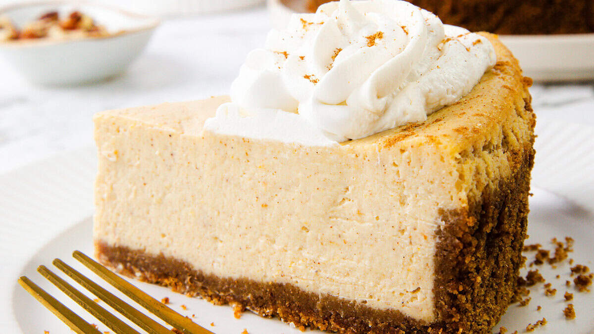 A slice of creamy cheesecake with a brown graham cracker crust, topped with a swirl of whipped cream and a sprinkle of cinnamon, sits on a white plate with a gold fork beside it—perfect for your Thanksgiving recipes collection.