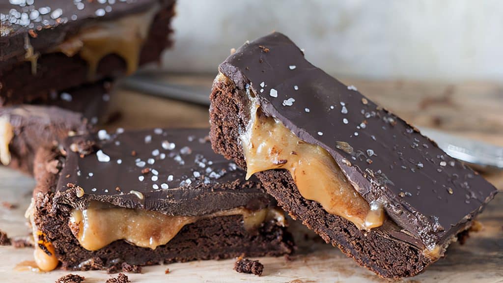 Two thick chocolate brownies with a gooey caramel layer in the center, topped with dark chocolate and a sprinkle of sea salt, are stacked on a wooden surface—the perfect treat for fans of Bar & Brownie Recipes.