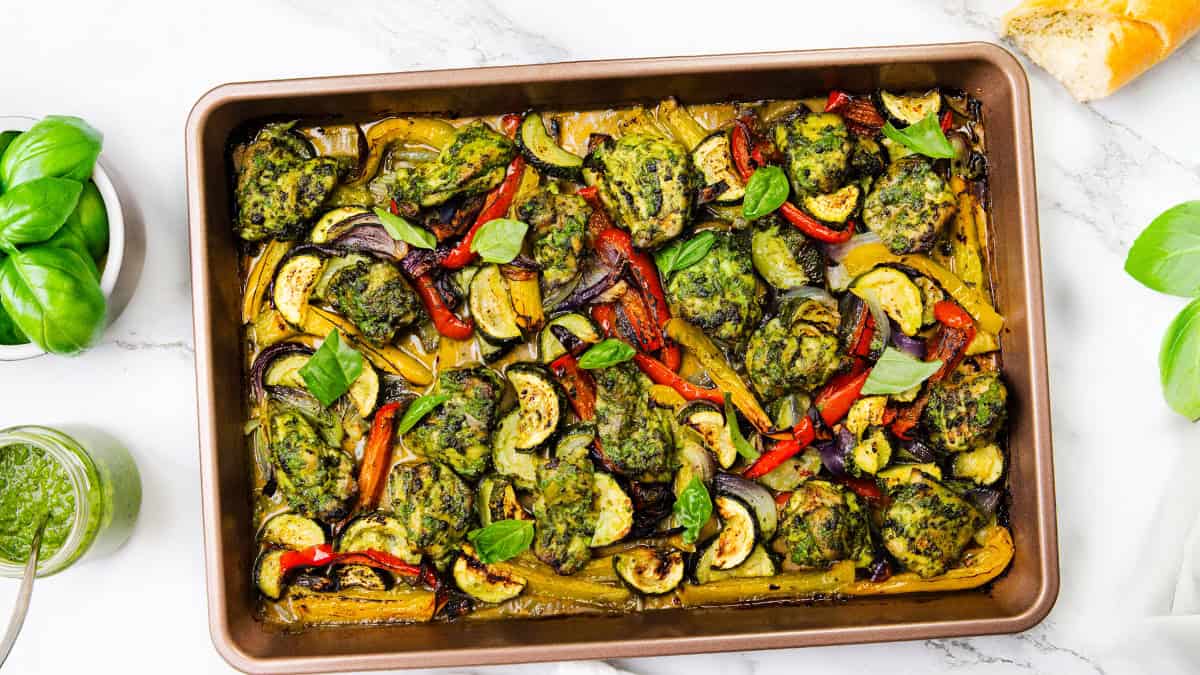 A baking pan filled with roasted vegetables and green herb-coated chicken pieces, garnished with fresh basil leaves, is featured on a white marble surface—perfect for fans of One Pot Recipes—alongside a jar of pesto and a baguette.