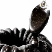 Sebuah sendok memercikkan glasir hitam mengkilap dengan bintik-bintik emas di atas kue hitam bergerigi yang dihiasi glasir keju krim hitam halus dengan latar belakang putih.