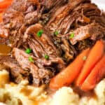 A plate containing shredded sirloin tip pot roast served on top of mashed potatoes, garnished with baby carrots and fresh parsley. The dish is accompanied by a savory brown gravy.