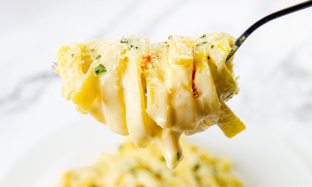 A close-up of a forkful of creamy fettuccine Alfredo pasta, garnished with grated Parmesan cheese and fresh herbs—an irresistible choice for easy dinners—held above a plate with more pasta in the background.