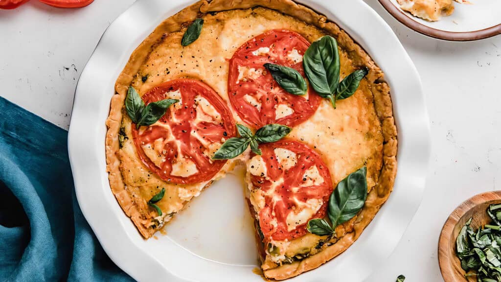 A homemade tomato and basil quiche with two slices missing, topped with fresh basil leaves, displayed on a white plate. The background includes a blue napkin and small wooden bowl with chopped herbs.