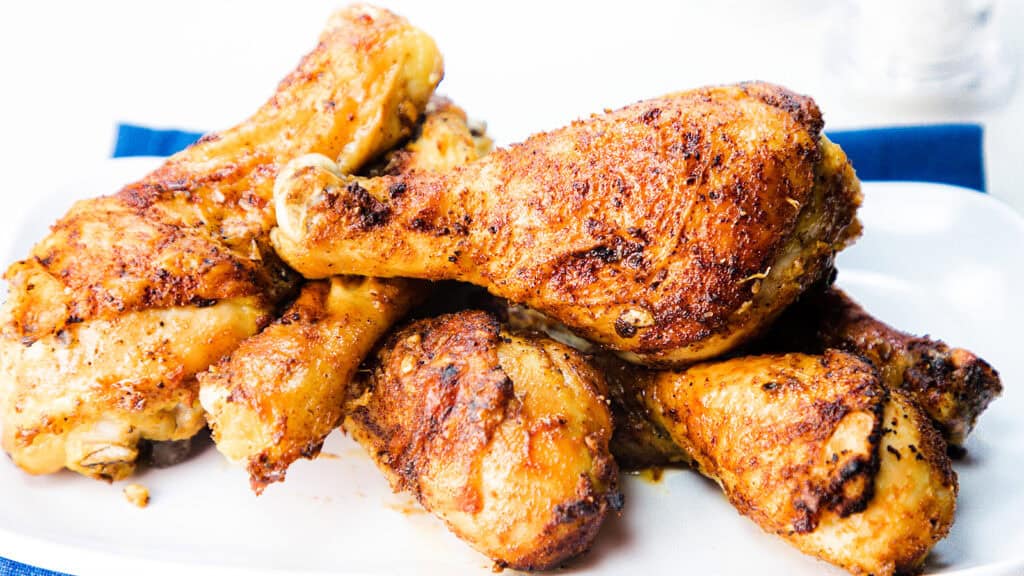 Sepiring stik ayam goreng berwarna cokelat keemasan yang ditumpuk tinggi, dibumbui dengan rempah-rempah dan rempah-rempah, di atas piring putih.