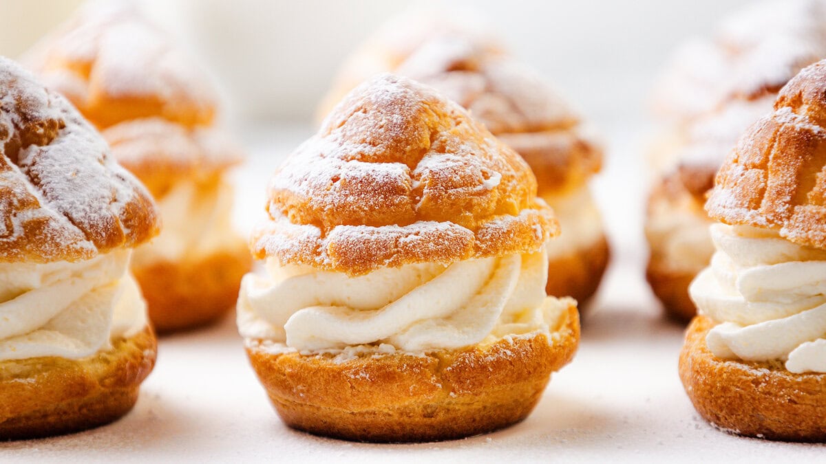 Golden cream puffs filled with swirled whipped cream and dusted with powdered sugar are arranged closely together on a light surface—an elegant addition to your Thanksgiving recipes. The background is softly blurred.